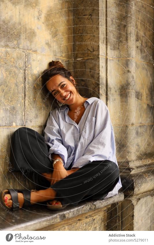 Lächelnde junge Frau sitzt auf einer Steinbank in der Nähe eines alten Gebäudes und schaut in die Kamera verträumt ruhen heiter allein sich[Akk] entspannen