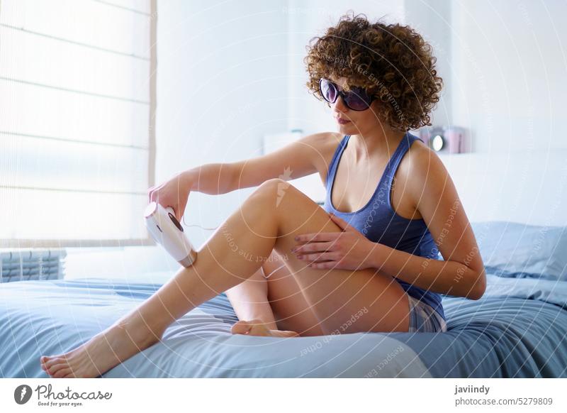 Seriöse Frau bei der Laser-Haarentfernung am Bein auf dem Bett mit Schutzbrille ipl Laser mit gepulstem Licht entfernen Behaarung schützend Schlafzimmer
