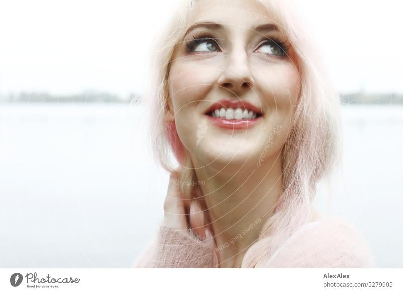 Junge Frau mit pink-blonden Haaren schaut in den Himmel und lächelt vor einem See mit Seeufer in der Ferne junge Frau Portrait schön schlank lange Haare
