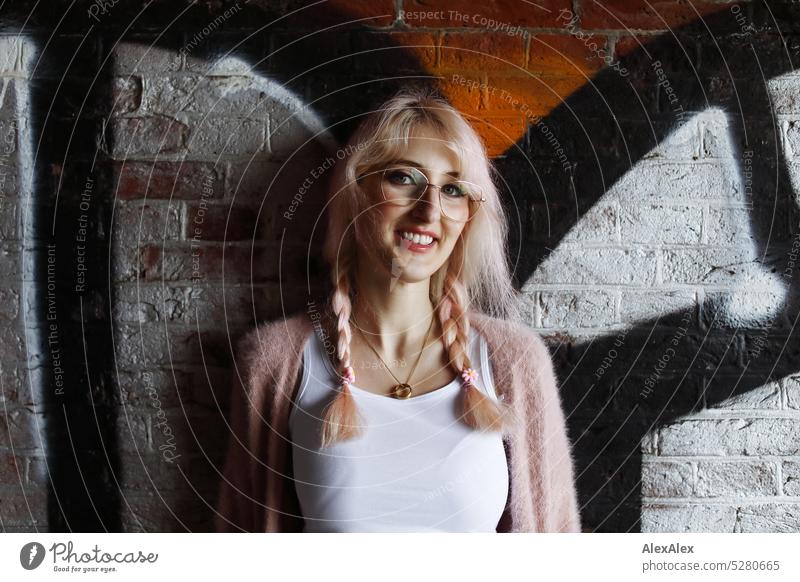 Junge Frau mit pink-blonden Haaren steht lächelnd vor einer Backsteinwand, die mit Graffiti verschandelt ist junge Frau Wand Portrait Brille schön schlank