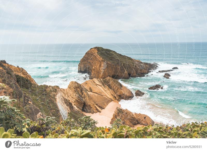 Steile Klippen, die in den gelb-orangen Sand eintauchen, der von den Wellen des Atlantiks in der Region Odemira, Portugal, bewässert wird. Auf den Spuren des Fischerpfads, Rota Vicentina
