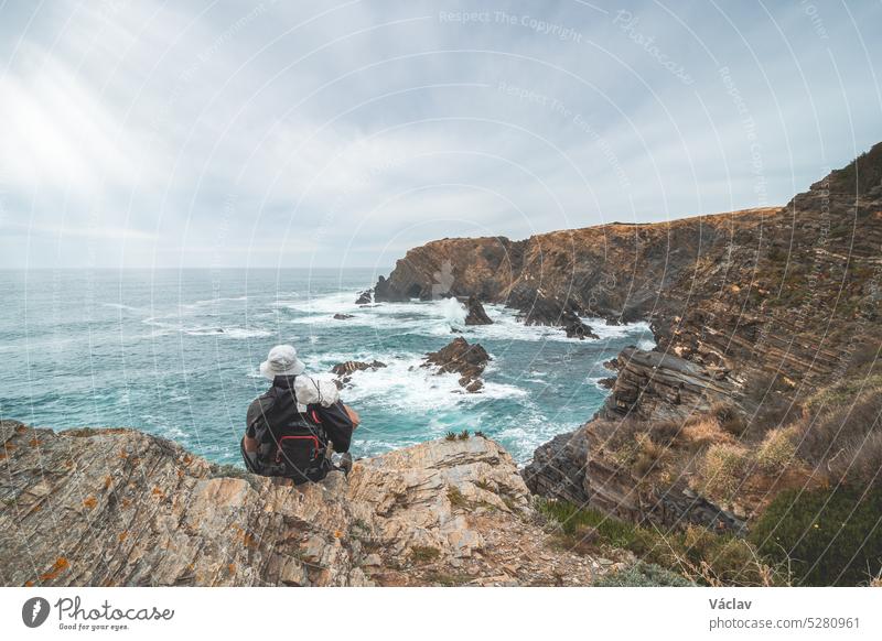 Wanderer und Abenteurer sitzen an einer zerklüfteten Fels- und Klippenküste am Atlantik in der Nähe der Stadt Odeceixe im Westen Portugals in der berühmten Touristenregion der Algarve. Sonnenuntergang