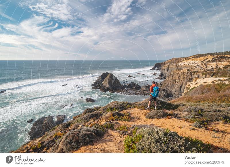 Freudiger Gesichtsausdruck eines jungen Rucksacktouristen, der am Rande einer Klippe mit Blick auf den Atlantischen Ozean in der Region Odemira im Westen Portugals steht. Wanderung entlang des Fisherman Trail, Rota Vicentina