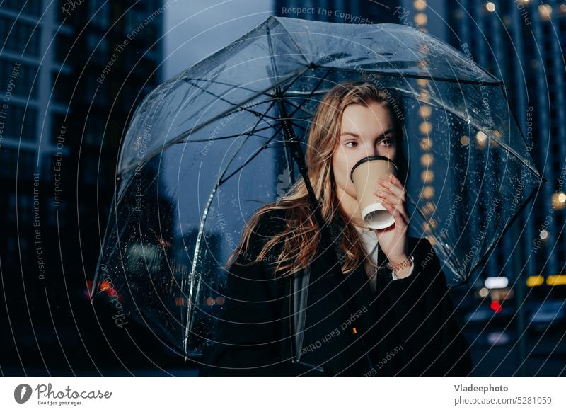 Blonde Frau im Mantel geht unter Regenschirm und hält Pappbecher in der Stadt Tasse Beteiligung Stehen Papier Wetter herbstlich Nacht kalt Kaffee unten