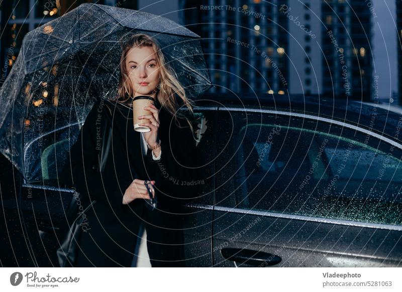 Eine Frau steht an einem regnerischen Abend unter einem Regenschirm neben ihrem Auto. Sie trinkt Kaffee zum Mitnehmen PKW jung Dame Nacht warten Taxi Miete