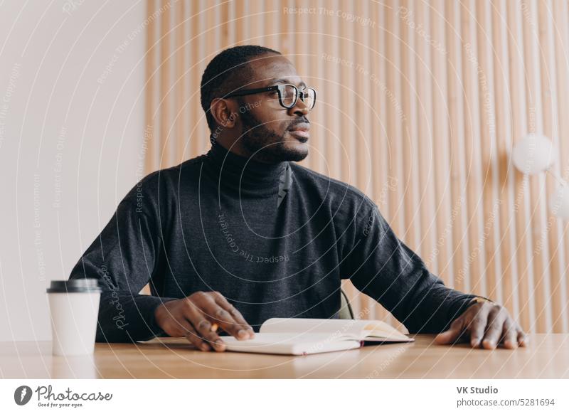 Nachdenklicher afrikanischer Geschäftsmann sitzt mit Stift und Agenda am Schreibtisch und schaut mit nachdenklichem Gesichtsausdruck weg jung Afrikanisch
