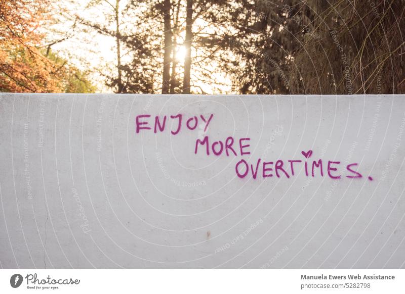 Hinter einer Betonmauer geht die Sonne auf. Auf der Mauer steht: "Enjoy more overtimes." betonmauer spruch überwinden überstunden arbeit stress gesellschaft