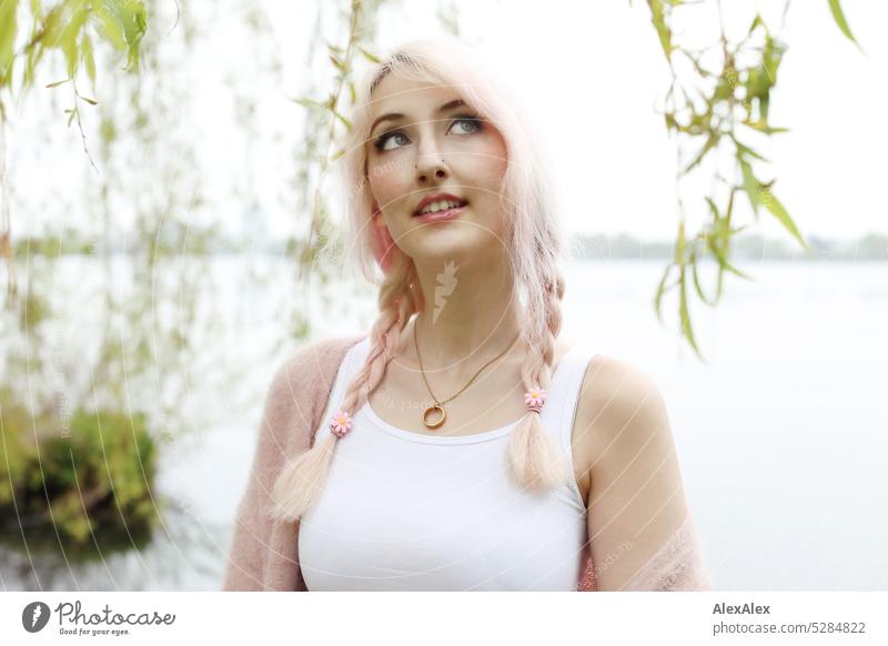 Junge Frau mit pink-blonden Haaren schaut in den Himmel und lächelt vor einem See mit Seeufer in der Ferne junge Frau Portrait schön schlank lange Haare