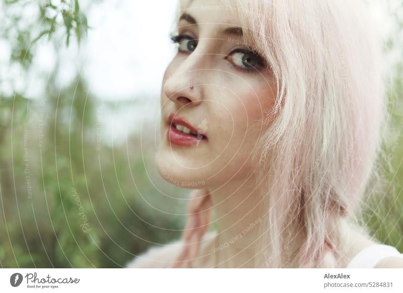 Junge Frau mit pink-blonden Haaren schaut seitlich in die Kamera und steht vor einem See mit Bäumen und Sträuchern am Ufer junge Frau Portrait schön schlank