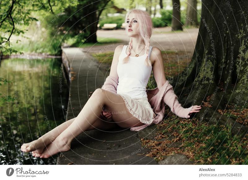 Junge Frau mit pink-blonden Haaren und langen Beinen sitzt am befestigten Ufer eines Sees unter einem Baum und schaut über den See junge Frau Portrait schön