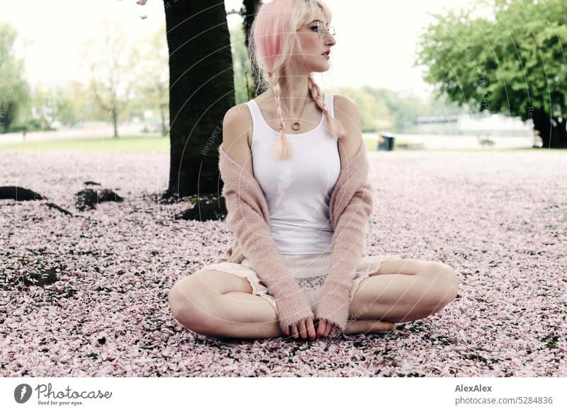 Junge Frau mit pink-blonden Haaren sitzt barfuß im Schneidersitz auf dem Boden eines Parks, der mit rosa Kirschblütenblättern bedeckt ist junge Frau Portrait