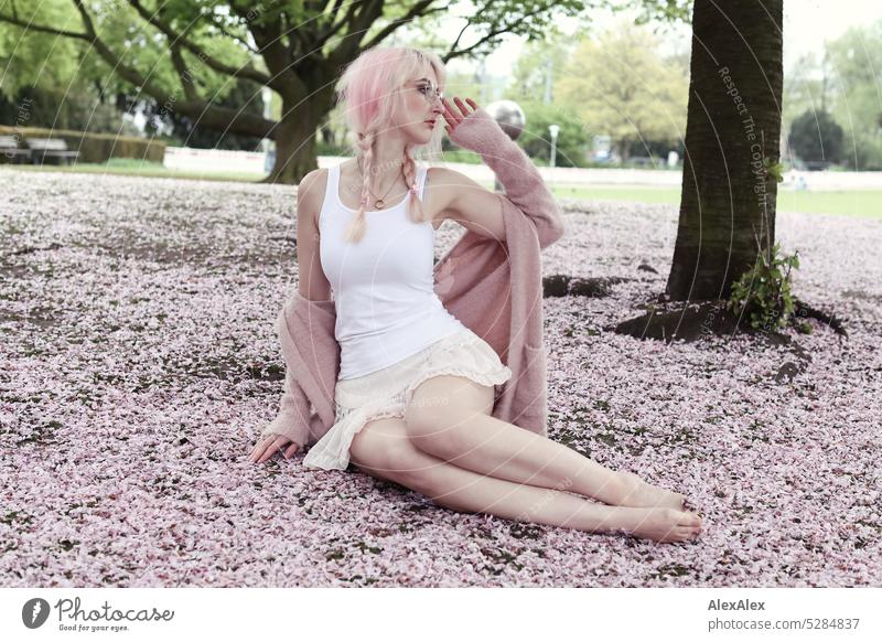 Junge Frau mit pink-blonden Haaren sitzt barfuß auf dem Boden eines Parks, der mit rosa Kirschblütenblättern bedeckt ist junge Frau Portrait schön schlank