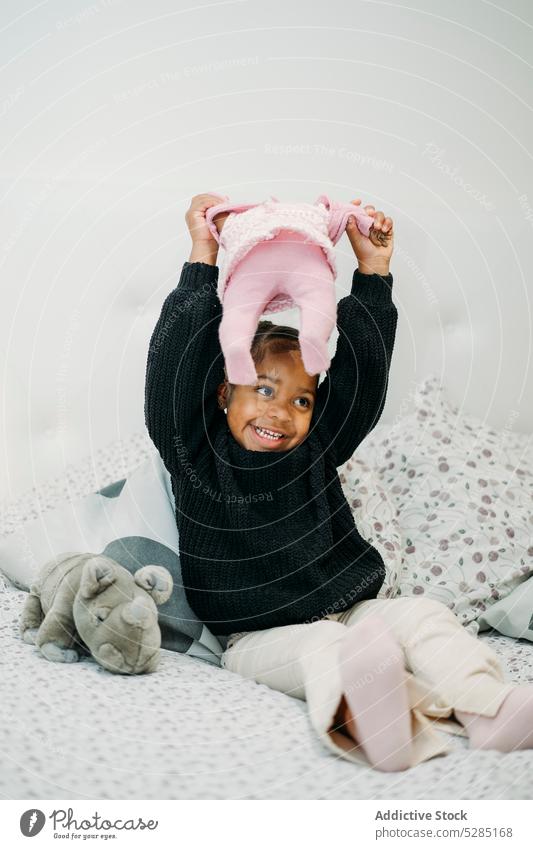 Glückliches kleines schwarzes Mädchen sitzt auf dem Bett und spielt mit Spielzeug Puppe Aktivität spielen Spaß Freude Spaß haben unterhalten Lächeln Kind