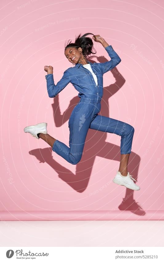 Glückliche schwarze Frau springt hoch auf rosa Hintergrund springen positiv Spaß haben heiter aufgeregt trendy Lächeln Stil Arme hochgezogen jung Afroamerikaner