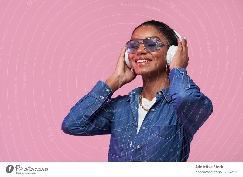 Lächelnde schwarze Frau mit Kopfhörern hört Musik auf rosa Hintergrund trendy Stil zuhören Mode Outfit Jeansstoff Sonnenbrille Afroamerikaner jung ethnisch