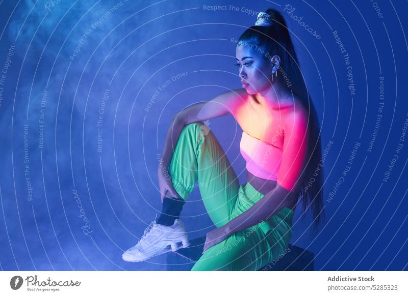Fit gemischte Rasse Frau sitzt auf Würfel in Rauch Model Projektor Körperhaltung Studioaufnahme Vorschein neonfarbig leuchten Dame jung ethnisch Outfit