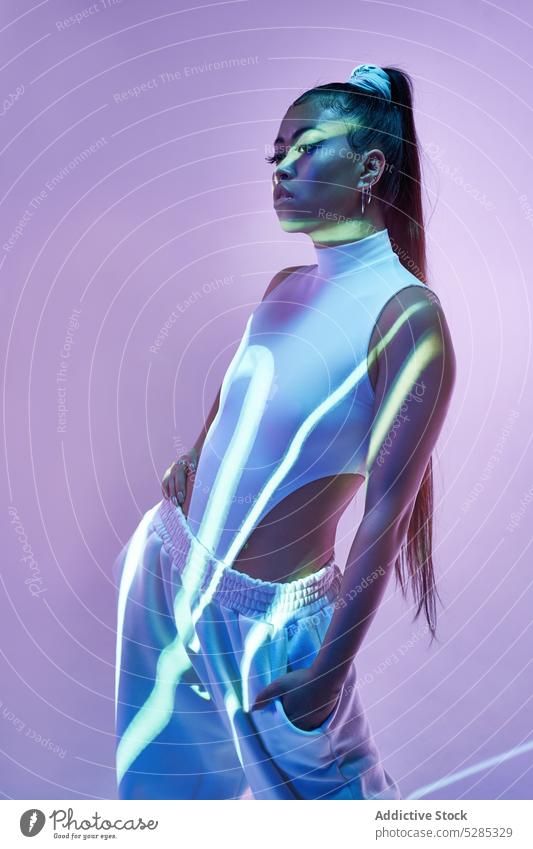 Gemischtrassige Frau im Studio mit Neonbeleuchtung und Linien aus dem Projektor Model Hand auf der Taille Hand-in-Tasche Körperhaltung emotionslos