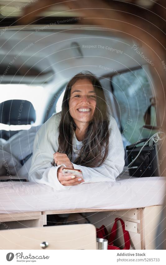 Positive junge ethnische Frau mit Smartphone und Lächeln im Wohnmobil Kleintransporter Lügen heiter Autoreise Reisender positiv benutzend SMS Urlaub Glück