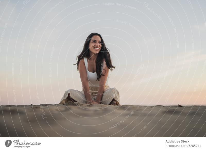 Unbeschwerte junge ethnische Frau am Strand sitzend bei Sonnenuntergang Lächeln Sand Feiertag Beine gekreuzt Kälte Tourist Glück positiv Sommer Urlaub