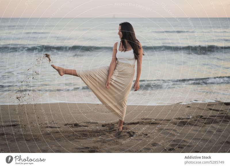 Unbekümmerte junge hispanische Frau, die am Meeresstrand in den Sand tritt Strand Bein angehoben Lächeln genießen Urlaub Sonnenuntergang Tourist Glück MEER