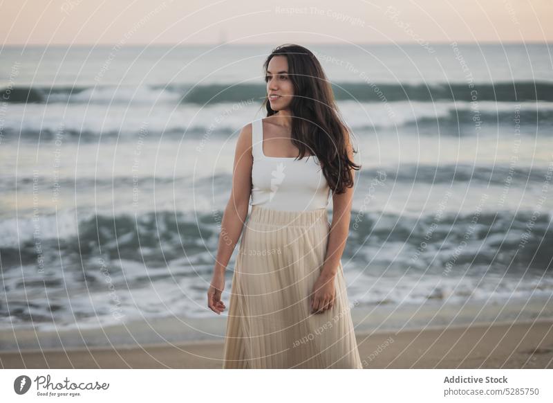 Fröhliche junge ethnische Frau, die im Sommerurlaub am sandigen Meeresufer steht Spaziergang Strand MEER genießen Feiertag Sonnenuntergang Reisender Sand Natur
