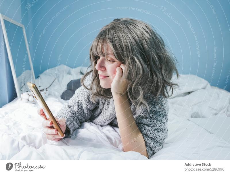 Lächelnde Frau mit Smartphone auf dem Bett positiv benutzend Browsen Gerät Schlafzimmer ruhen heimwärts Spiegel Mobile Komfort Apparatur im Inneren Surfen Lügen