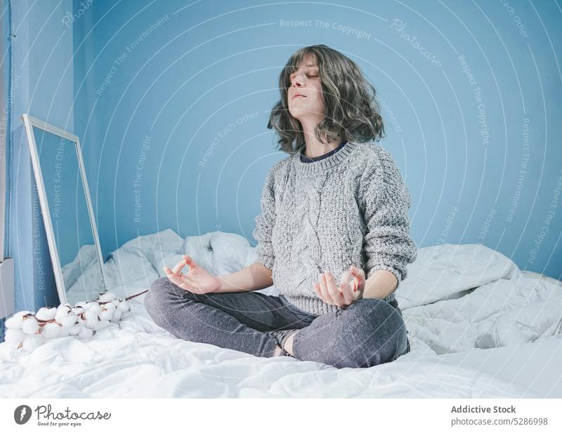 Frau im Pullover meditiert auf dem Bett meditieren Yoga Lotus-Pose Augen geschlossen üben Zen Schlafzimmer warme Kleidung Beine gekreuzt Wand Windstille Komfort