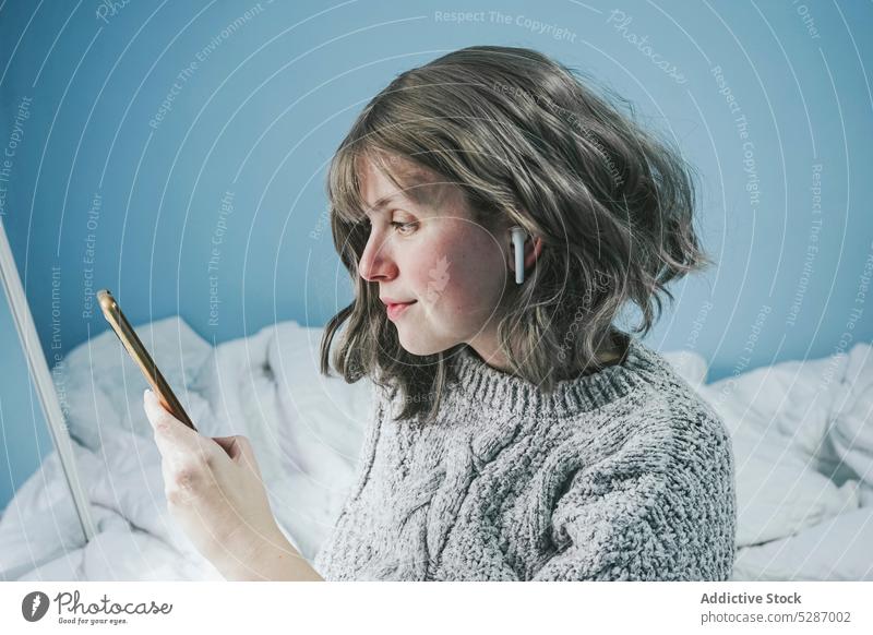 Lächelnde Frau mit Smartphone auf dem Bett positiv benutzend Ohrstöpsel Browsen zuhören Gerät Schlafzimmer ruhen heimwärts soziale Netzwerke Mobile Komfort