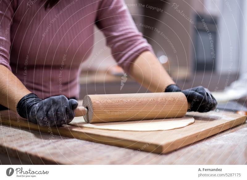 Detail einer Frau, die einen Pizzateig mit einem hölzernen Nudelholz auf einem Küchentisch knetet und dabei schwarze Handschuhe trägt Kneten Pizzabäcker
