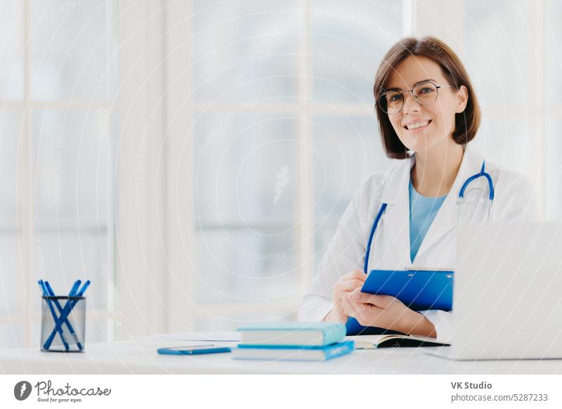 Eine Ärztin schreibt ein Rezept auf ein spezielles Formular, arbeitet in einer Privatklinik, trägt einen weißen Arztkittel, ist bereit, Patienten zu empfangen, posiert am Arbeitsplatz. Lächelnder Arzt oder medizinischer Arbeiter hält Klemmbrett