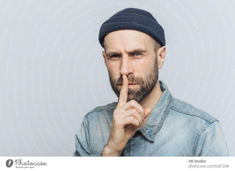 Seriöser, angenehm aussehender, bärtiger Mann mit geheimnisvollem Gesichtsausdruck, der den Zeigefinger auf die Lippen legt und darum bittet, persönliche Informationen vertraulich zu behandeln, trägt einen stilvollen Hut und ein Jeanshemd, isoliert auf einer grauen Wand