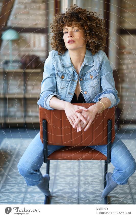 Ernste junge Frau in Jeansjacke auf einem Lederstuhl sitzend ernst Haare berühren emotionslos krause Haare Dessous Jeansstoff Jeanshose Vorschein Stil Raum
