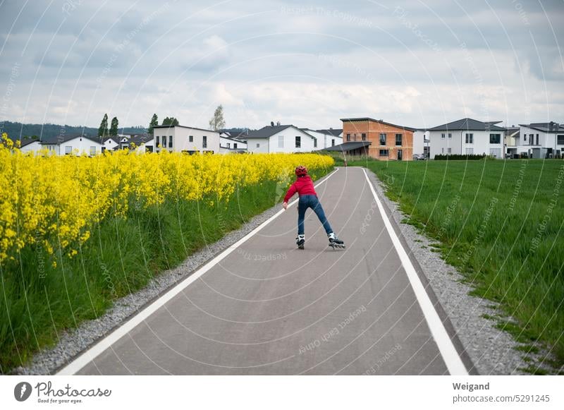 Ein Kind fährt auf einem frisch asphaltierten Weg zwischen Wiesen und Rapsfeldern Inlineskates in Richtung einer Neubausiedling Sport Rollerblades Straße