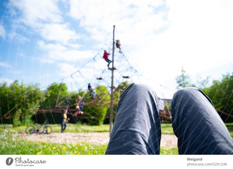 Beine eines Mannes, der bei schönem Wetter auf einer Wiese liegt und seinen Kindern im Hintergrund beim Spielen und Klettern zusieht, während er ausruht Pause
