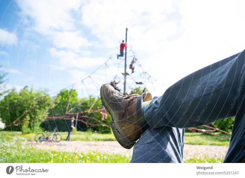 Überkreuzte Beine eines Mannes, der bei schönem Wetter auf einer Wiese liegt und seinen Kindern im Hintergrund beim Spielen und Klettern zusieht, während er ausruht