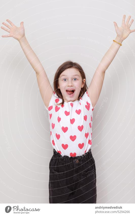Nette lächelnde Kind Mädchen in weißem T-Shirt mit roten Herzen und stilvolle schwarze Jeans hebt fröhlich seine Hände auf weißem Hintergrund. valentines day