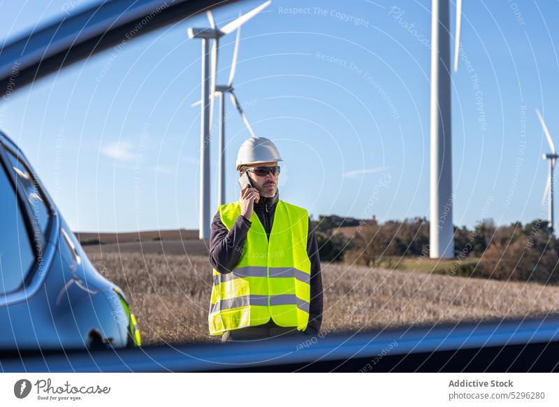Seriöser männlicher Arbeiter, der in der Nähe eines Autos mit seinem Smartphone spricht Mann benutzend Telefonanruf Fenster PKW Weste Schutzhelm Fahrzeug Wind