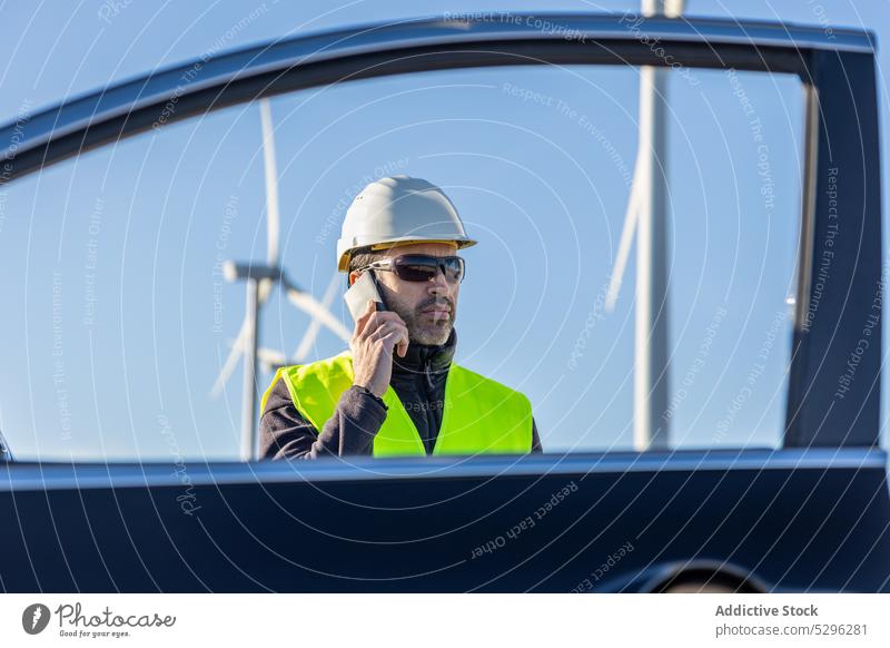 Seriöser männlicher Arbeiter, der in der Nähe eines Autos mit seinem Smartphone spricht Mann benutzend Telefonanruf Fenster PKW Weste Schutzhelm Fahrzeug Wind