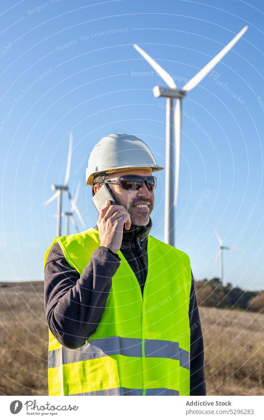 Ingenieur mit Schutzhelm spricht per Smartphone gegen Windkraftanlagen Mann Landschaft Fahrbahn Arbeiter benutzend Telefonanruf Turbine Öko alternativ Energie