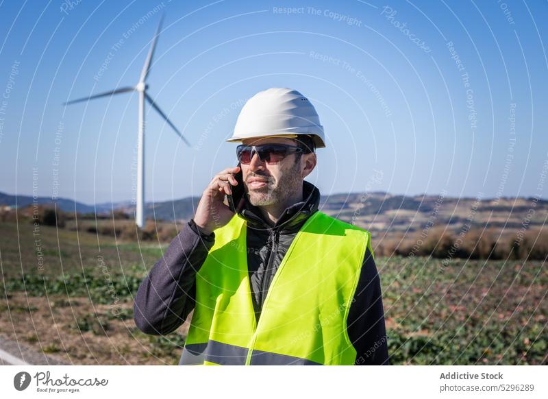 Ingenieur mit Schutzhelm spricht per Smartphone gegen Windkraftanlagen Mann Landschaft Fahrbahn Arbeiter benutzend Telefonanruf Turbine Öko alternativ Energie