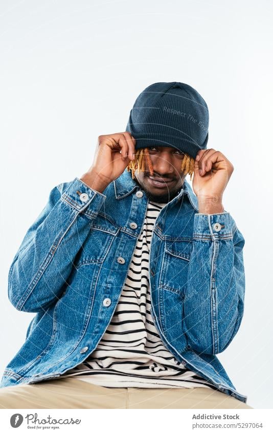 Stilvoller schwarzer Mann, der seinen Hut auf die Stirn zieht positiv Streetstyle Vorschein Jeansstoff spielerisch ziehen Studioaufnahme Rastalocken