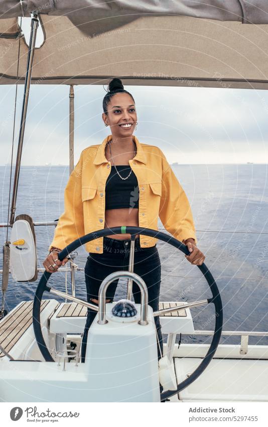 Fröhliche schwarze Frau steht am Steuerrad einer Jacht MEER Lenkrad Lächeln Sommer Stil Ausflug reisen Segelboot Glück Afroamerikaner Jacke positiv Wasser
