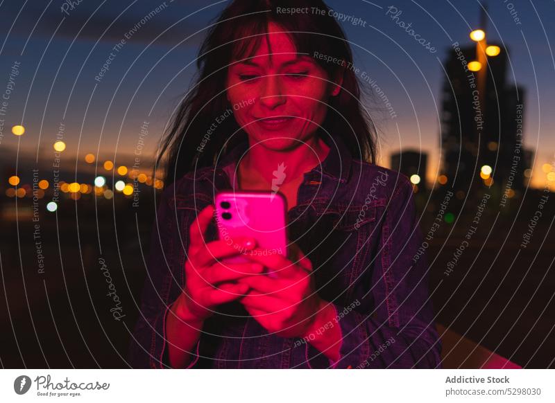Lächelnde Frau mit Smartphone im Neonlicht auf der Straße benutzend Nacht Großstadt heiter Glück neonfarbig Browsen urban leuchten Apparatur Mobile Gerät dunkel