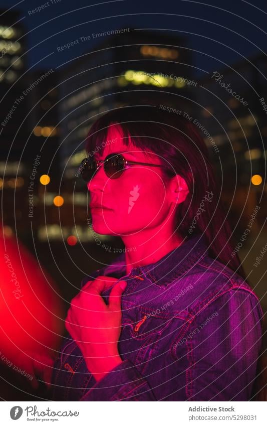 Ruhige Frau mit Brille im Neonlicht auf der Straße Nacht neonfarbig verträumt Großstadt leuchten Gebäude Nachtleben Licht Stil urban dunkel Jeansstoff
