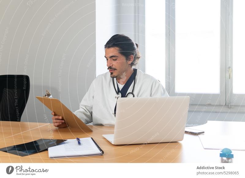 Konzentrierter Arzt prüft Informationen auf dem Klemmbrett Mann Röntgenbild Laptop Zwischenablage Krankenhaus Klinik Sanitäter ethnisch Arbeit jung