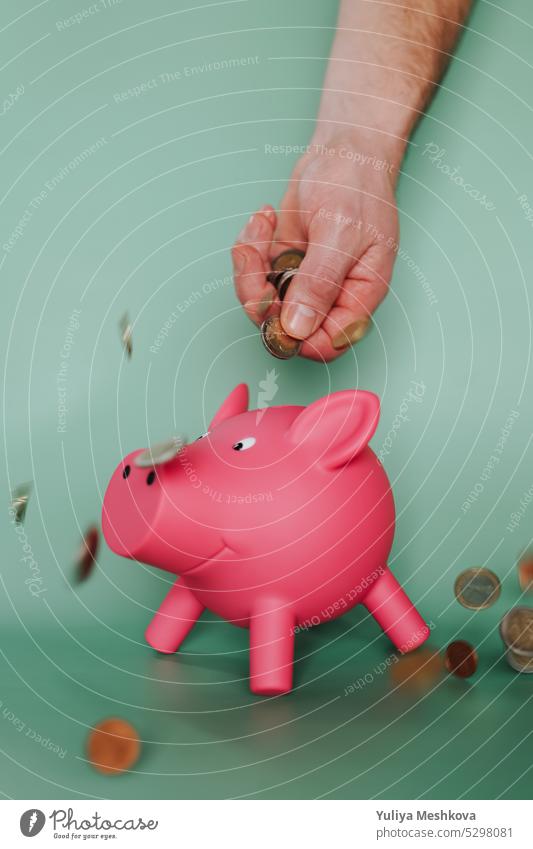 Fallende Euro-Münzen aus einer männlichen Hand und ein rosa Sparschwein auf einem grünen Hintergrund. Schweinchen Bank Einsparungen Ausgaben Konzept fallend