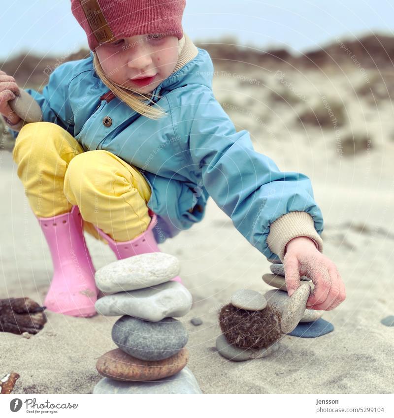 Ein kleines Mädchen spielt mit Steinen am Strand bunt pink gelb hellblau rosa Gummistiefel Kind Kindheit kindlich Spielen Außenaufnahme Farbfoto Freude