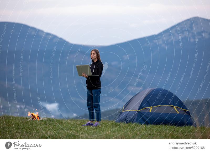 Junge Freiberuflerin arbeitet am Laptop in den Bergen am Abend. Touristisches Mädchen sitzt am Lagerfeuer und hat Spaß. Raum kopieren. jung Frau Sitzen online