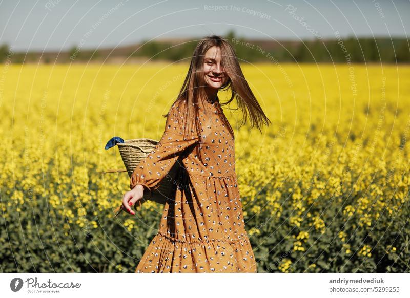 schöne junge Frau geht in einem Feld von gelben Raps. Mädchen brunette lange Haare fliegen im Wind in Kleid und Strohhut gekleidet. Sommerurlaub Konzept Natur