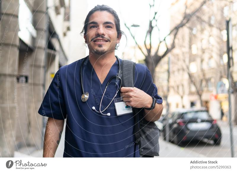 Fröhlicher ethnischer Arzt mit Rucksack auf der Straße Mann Großstadt Stethoskop Lächeln positiv medizinisch Klinik Gesundheitswesen männlich Uniform Glück